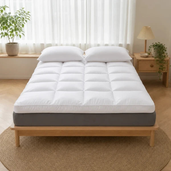 Surmatelas épais matelassé blanc posé sur un lit, matelas gris visible, deux oreillers