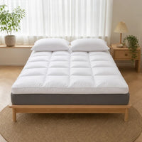 Surmatelas épais matelassé blanc posé sur un lit, matelas gris visible, deux oreillers