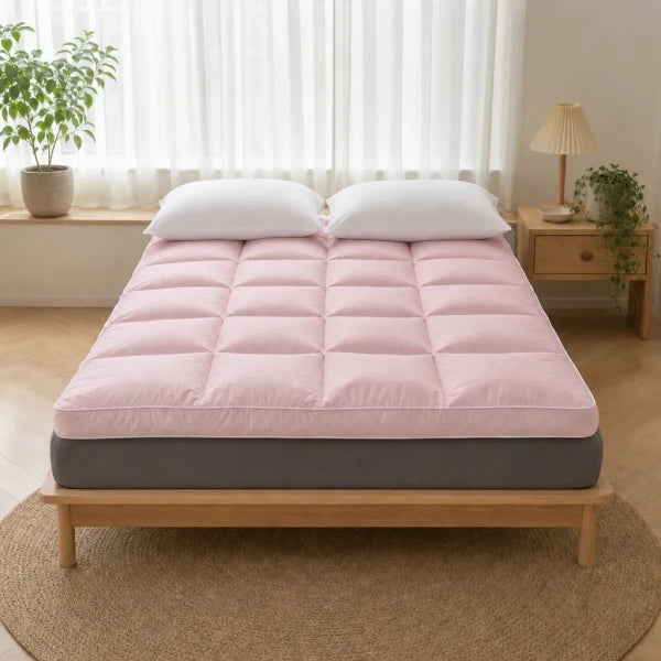 Surmatelas épais matelassé rose posé sur un lit, matelas gris visible, deux oreillers blancs