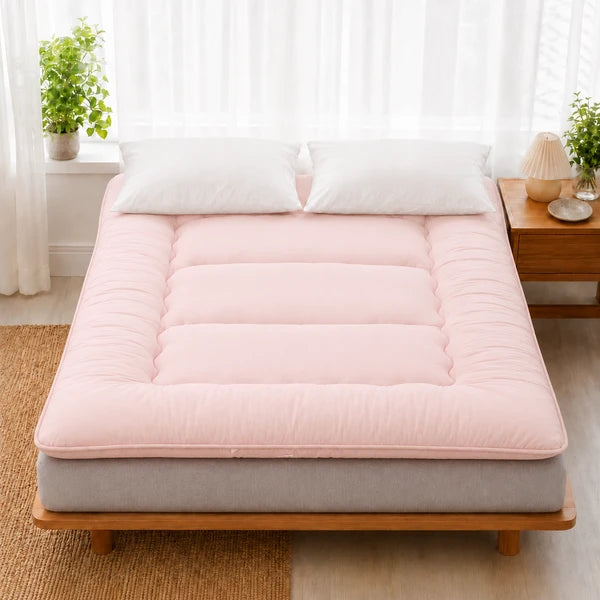 Surmatelas ouate rose pose sur un lit en bois clair, deux oreillers blancs