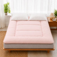 Surmatelas ouate rose pose sur un lit en bois clair, deux oreillers blancs