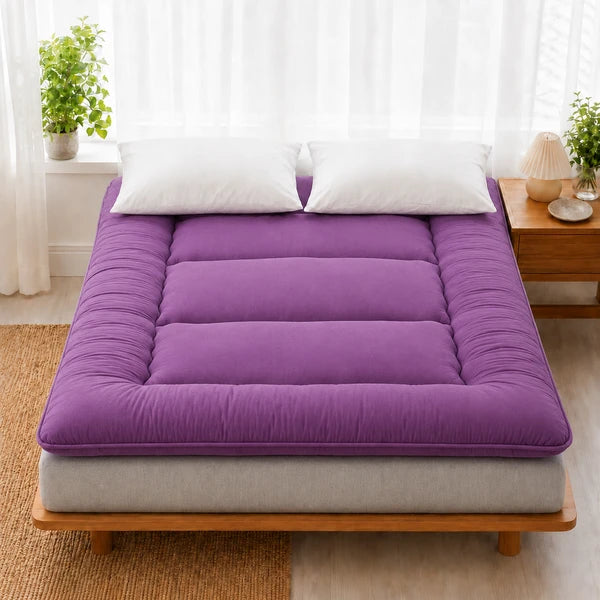 Surmatelas ouate violet pose sur un lit en bois clair, deux oreillers blancs
